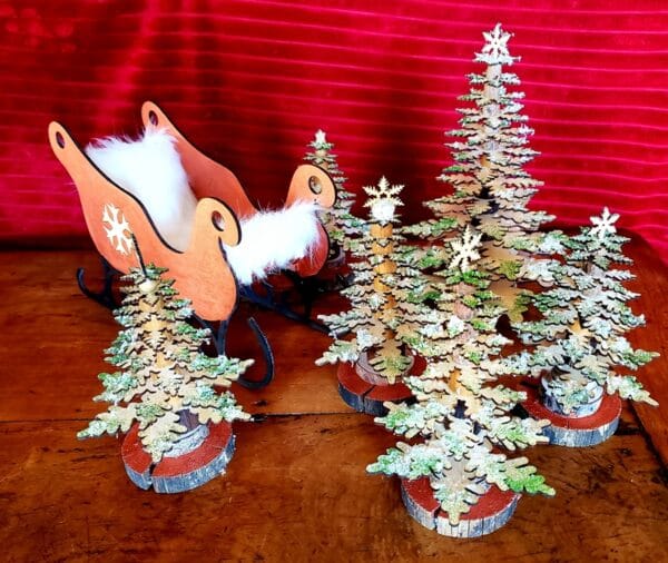 Un traîneau rouge en bois avec une doublure en fourrure blanche se trouve à côté de plusieurs petits arbres de Noël décoratifs en bois superposés sur une surface en bois, avec un arrière-plan rouge et texturé.