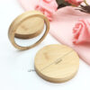 Taille-Miroir-de-Poche-en-Bois Un miroir compact rond en bambou, d'une largeur de 72 mm et d'une épaisseur de 12 mm, est présenté ouvert à côté d'un tissu rose et de roses artificielles roses. Un côté montre le miroir, l'autre le couvercle en bambou.