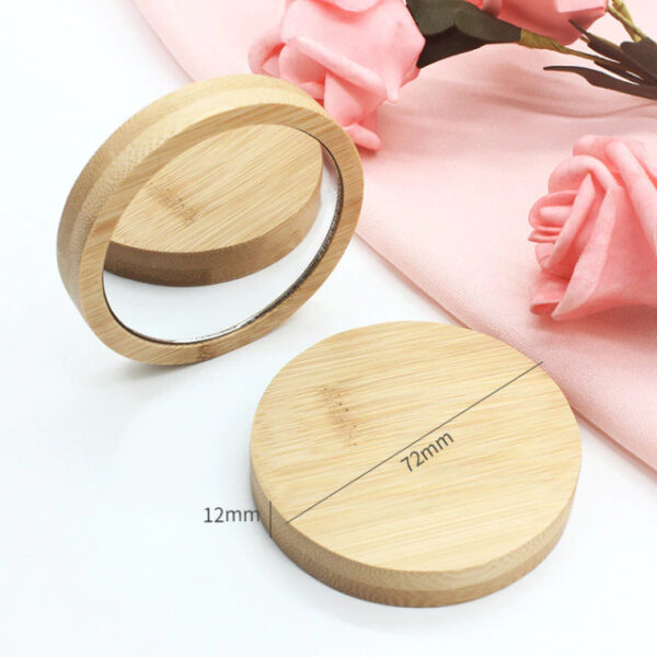 Un miroir compact rond en bambou, d'une largeur de 72 mm et d'une épaisseur de 12 mm, est présenté ouvert à côté d'un tissu rose et de roses artificielles roses. Un côté montre le miroir, l'autre le couvercle en bambou.