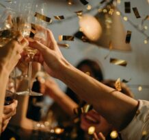Un groupe de personnes porte un toast de célébration au milieu de confettis dorés et de guirlandes lumineuses, capturant l'esprit joyeux des fêtes dans une atmosphère festive. {{brizy_dc_image_alt imageSrc=