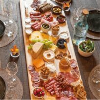 Une longue planche en bois remplie de fromages assortis, de charcuterie, de pain, de raisins, d'olives, de pâtes à tartiner, de noix et de miel, entourée d'assiettes vides, de verres et d'une bouteille d'eau sur une table en bois rustique. {{brizy_dc_image_alt imageSrc=